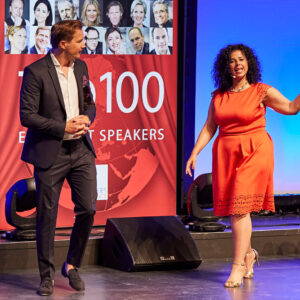 Due relatori sul palco di un evento, con un pannello che celebra i 100 migliori speaker del mondo.