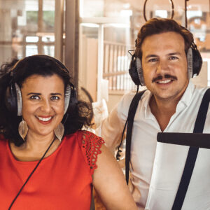 Due presentatori sorridenti in studio radiofonico, entrambi con cuffie, trasmettono energia e convivialità.