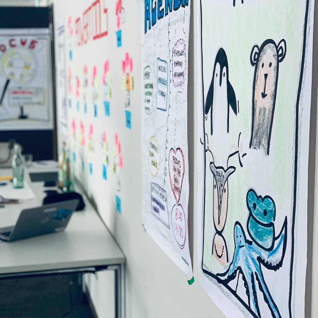 Colorful sketches of animals and an agenda displayed on a wall in a collaborative meeting space with a table and laptop.