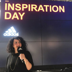 Evento Inspirational Day di Adidas con presentatrice al microfono sullo sfondo di un pannello digitale.