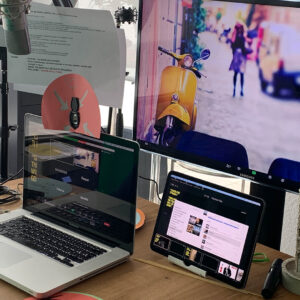 Postazione di lavoro con laptop, tablet e microfono, utilizzata per una videoconferenza con sfondo di una vespa gialla.