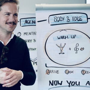 Uomo sorridente presenta un poster su tecniche di riscaldamento per il corpo e la voce durante un workshop.