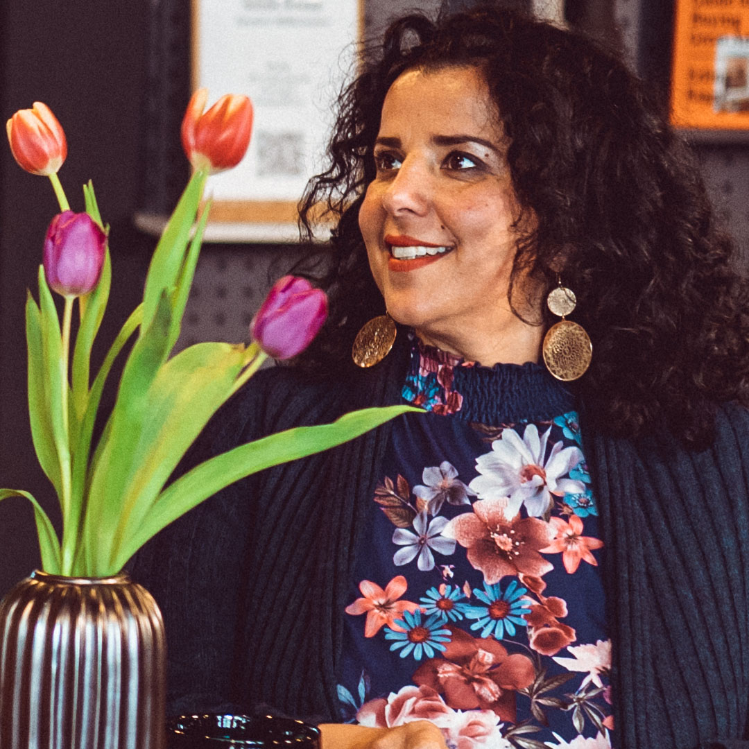 Eine lächelnde Frau mit lockigem Haar sitzt neben einer Vase mit bunten Tulpen und trägt auffällige Ohrringe.