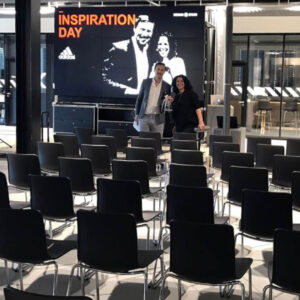 Two people stand in a seated auditorium with a display screen announcing Inspiration Day at an Adidas event.